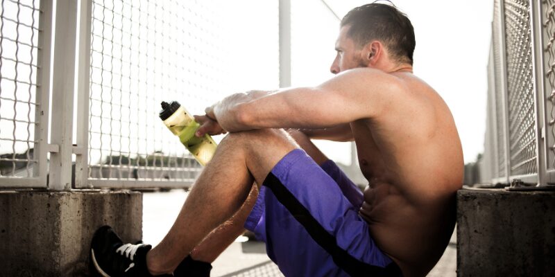 Athletic man sitting after workout holding water bottle, illustrating post-exercise fatigue and muscle recovery needs